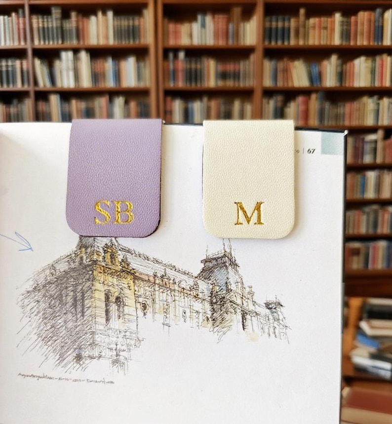 May include: Two leather magnetic bookmarks. One is lavender with gold embroidered initials "SB", the other is cream with gold embroidered initial "M". The bookmarks are on a book with a sketch of a building.