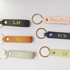 Puede incluir: Una colección de llaveros personalizados de cuero en varios colores, incluyendo negro, marrón, rosa, azul marino y verde claro. Cada llavero presenta herrajes dorados e iniciales o un nombre en relieve. Los llaveros están dispuestos sobre una superficie blanca.