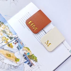 May include: Two leather bookmarks, one tan and one cream, rest on an open book. The tan bookmark has gold embroidered initials "HM", while the cream bookmark displays the letter "M". The book features sketches and text.