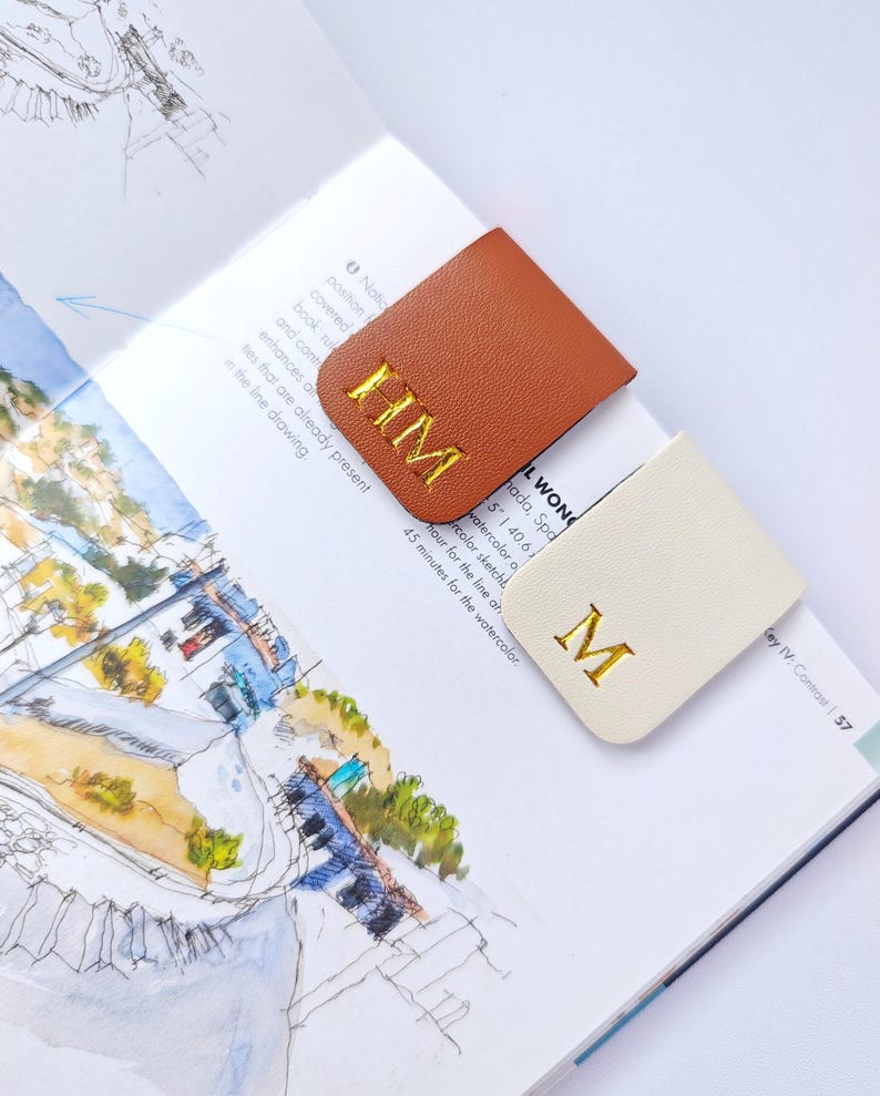 May include: Two leather bookmarks, one tan and one cream, rest on an open book. The tan bookmark has gold embroidered initials "HM", while the cream bookmark displays the letter "M". The book features sketches and text.