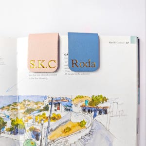 May include: Two leather bookmarks, one light pink and one blue, rest on a book page. The pink bookmark has the gold letters "S.K.C", and the blue one has "Roda". The book is open to a watercolor illustration.