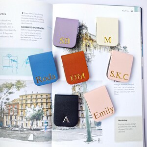 May include: A collection of personalized leather book page holders in various colors, including lavender, blue, orange, black, cream, and pink. Each holder features gold lettering with initials or names. The items are displayed on an open book.