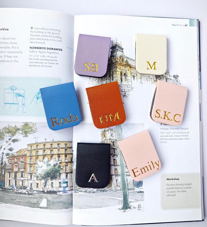 May include: A collection of personalized leather book page holders in various colors, including lavender, blue, orange, black, cream, and pink. Each holder features gold lettering with initials or names. The items are displayed on an open book.