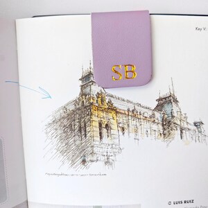 May include: A lavender leather bookmark with gold embroidered initials "SB" is clipped to a book page with a detailed architectural sketch. The bookmark is rectangular with a curved top.