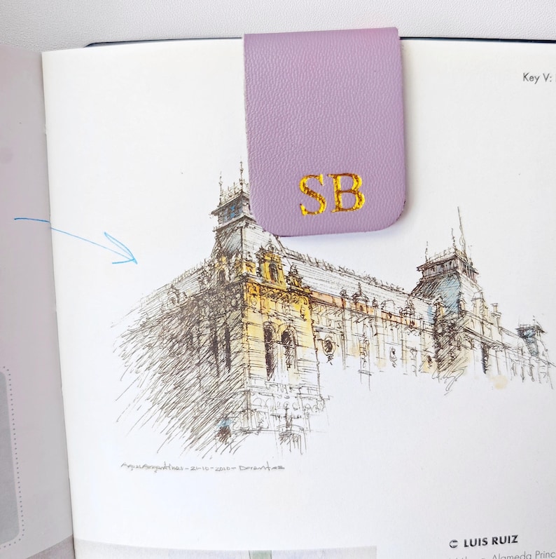 May include: A lavender leather bookmark with gold embroidered initials "SB" is clipped to a book page with a detailed architectural sketch. The bookmark is rectangular with a curved top.