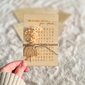Può includere: Una scheda puzzle in legno con il testo "EIN KLEINES RATSEL für dich" e una griglia di ricerca di parole. La scheda è decorata con fiori secchi, un arcobaleno di legno e spago. Un'idea regalo unica.