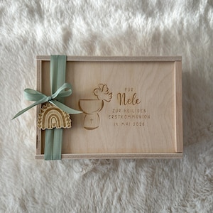 Puede incluir: Una caja de madera con una cinta verde claro y un adorno de arcoíris de madera. La caja está grabada con texto y un diseño de cáliz y paloma. El texto incluye un nombre y una fecha.