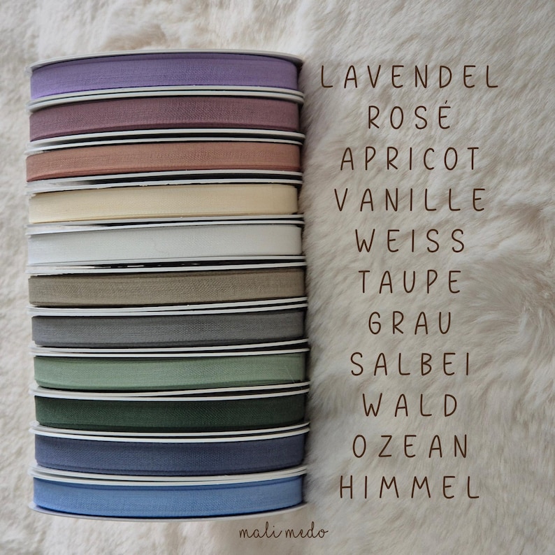 Puede incluir: Una pila de carretes de cinta de varios colores, incluyendo lavanda, rosa y azul. La imagen incluye texto con nombres de colores. La cinta est&aacute; sobre una superficie blanca texturizada.