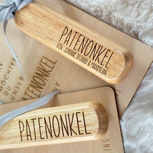 Może przedstawiać: Drewniane prostokątne zawieszki na prezenty ze słowem "PATENONKEL" wygrawerowanym pogrubioną czcionką. Jedna zawieszka zawiera imiona "VON JOHANNA, RICHARD & MAXIMILIAN". Zawieszki są związane szarą wstążką. Tło jest w jasnobrązowym kolorze.