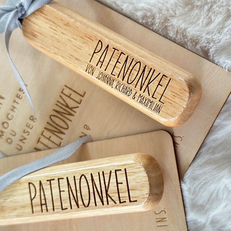 Może przedstawiać: Drewniane prostokątne zawieszki na prezenty ze słowem "PATENONKEL" wygrawerowanym pogrubioną czcionką. Jedna zawieszka zawiera imiona "VON JOHANNA, RICHARD & MAXIMILIAN". Zawieszki są związane szarą wstążką. Tło jest w jasnobrązowym kolorze.