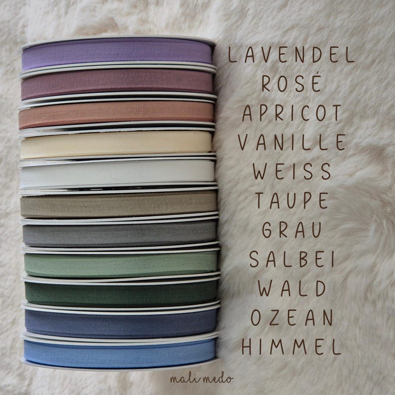 Puede incluir: Una pila de carretes de cinta de varios colores, incluyendo lavanda, rosa, albaricoque y azul. La imagen incluye texto con nombres de colores. La cinta est&aacute; sobre una superficie blanca texturizada.