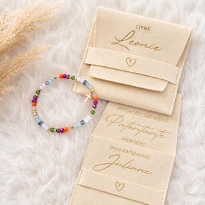 Può includere: Un braccialetto di perline con pietre multicolori e un piccolo ciondolo a croce dorata, accanto a due sacchetti di velluto beige. Il sacchetto superiore riporta il testo "LIEBE Leonie" con un cuore, quello inferiore "MÖCHTEST DU MEINE Patentante WERDEN? DEIN PATENKIND Juliane" con un cuore.