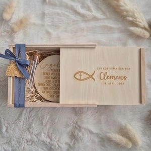 Pode incluir: Uma caixa deslizante de madeira clara contendo um disco de madeira com texto em alemão, um pequeno frasco de vidro e aparas de madeira. A tampa apresenta um símbolo de peixe com uma cruz, o texto ZUR KONFIRMATION VON Clemens e 28. APRIL 2026.
