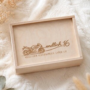 Könnte beinhalten: Eine hellfarbene Holzkiste mit Schiebedeckel. Der Deckel zeigt ein eingraviertes Motorrad-Design und den Text "endlich 16" sowie zusätzlichen Text. Die Kiste ist vor einem weichen, weißen Hintergrund platziert.