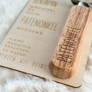 Może przedstawiać: Zestaw drewnianej tabliczki i otwieracza do butelek. Tabliczka ma wygrawerowany niemiecki tekst, w tym słowo "PATENONKEL". Otwieracz do butelek ma drewniany uchwyt z wygrawerowanym słowem "PATENONKEL".