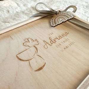 Puede incluir: Una caja de madera de color claro con una cinta y una etiqueta decorativa. La caja est&aacute; grabada con el nombre "Adriana", las palabras "ZUR TAUFE" y la fecha "11. Mai 2025". Tambi&eacute;n hay un dise&ntilde;o de paloma y c&aacute;liz grabado.