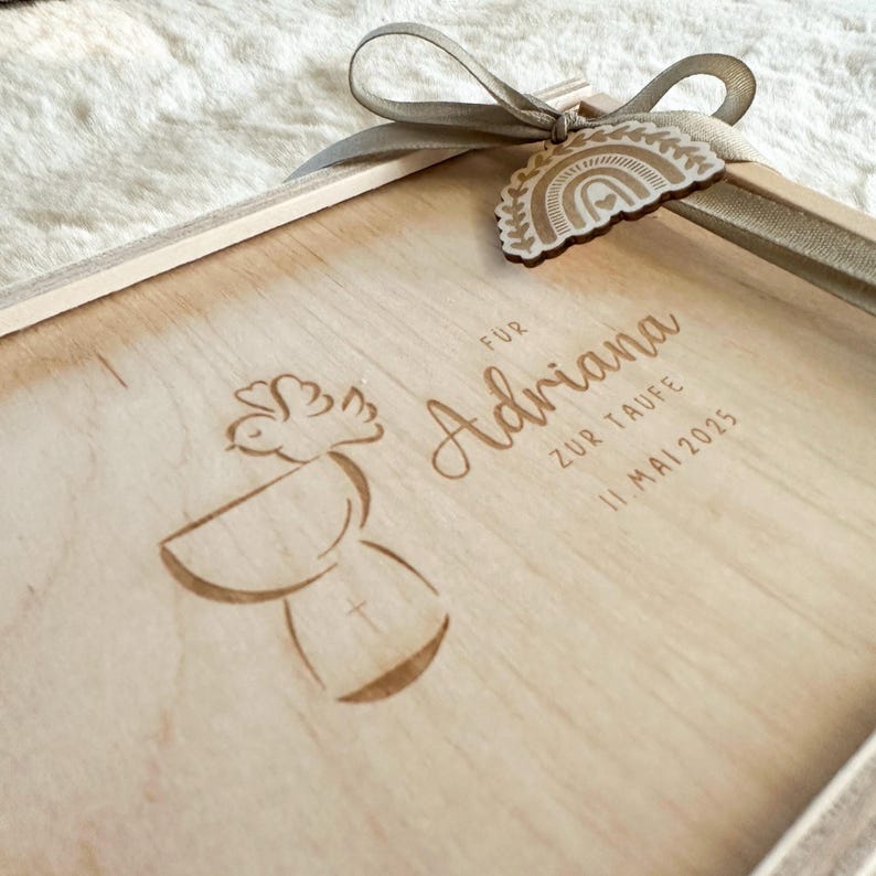 Puede incluir: Una caja de madera de color claro con una cinta y una etiqueta decorativa. La caja est&aacute; grabada con el nombre "Adriana", las palabras "ZUR TAUFE" y la fecha "11. Mai 2025". Tambi&eacute;n hay un dise&ntilde;o de paloma y c&aacute;liz grabado.