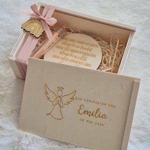 Puede incluir: Una caja de regalo de madera con una cinta rosa claro y una etiqueta de madera en forma de arcoíris. En el interior, un pequeño vial de vidrio, una placa redonda de madera con texto grabado y una tapa de caja de madera con una ilustración de ángel y el texto "ZUR KOMMUNION VON Emilia 09. MAI 2026".