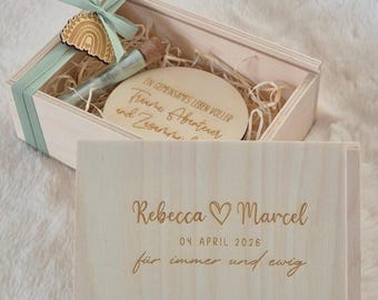 Op de afbeelding: Een lichtgekleurde houten geschenkdoos met een schuifdeksel gegraveerd met de namen Rebecca en Marcel, de datum 04. APRIL 2026 en de tekst für immer und ewig. Binnenin een ronde houten schijf, een glazen flesje en een regenboogvormig houten label.