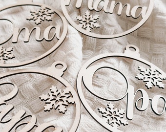 Könnte beinhalten: Vier hölzerne Weihnachtsschmuckstücke mit den Wörtern "Mama", "Papa", "Oma" und "Opa" in Schreibschrift. Jedes Ornament ist ein Kreis mit einem Schneeflocken-Design und einer kleinen Schlaufe zum Aufhängen. Die Ornamente sind hellbraun.