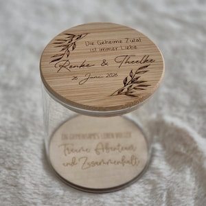May include: A clear glass jar with a round bamboo lid engraved with the text Die Geheime Zutat ist immer Liebe, Renke & Theelke, and 26. Juni 2026, accompanied by leaf illustrations. The base of the jar features an engraved wooden insert.