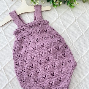 Puede incluir: Un pelele de bebé tejido de color lavanda con un patrón de hojas texturizadas. El pelele tiene tirantes y un escote con volantes. Se muestra en una percha de madera sobre un fondo blanco texturizado.