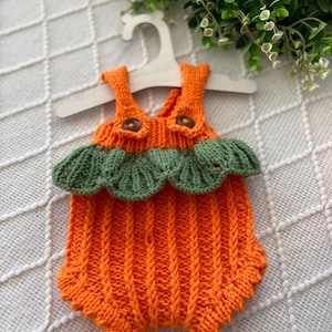 Peut inclure: Un barboteuse pour bébé tricotée orange, ornée de vert, évoquant une citrouille. Elle présente des boutons marron sur les bretelles et est suspendue à un cintre en bois. Le vêtement est fait d'un fil doux et texturé.