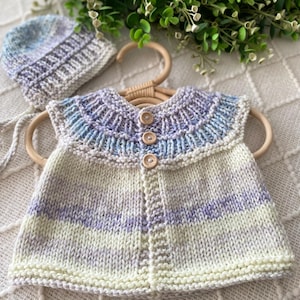 May include: A knitted baby outfit set, comprising a vest and matching hat. The vest features pale yellow, lavender, and blue hues, fastened with wooden buttons. The hat mirrors this colour scheme. The set is presented on a wooden hanger.