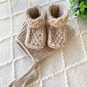 May include: A beige knitted baby set, including a pair of booties and a bonnet. The booties have a rolled cuff and a textured knit pattern. The bonnet has ties for securing. The set is made of soft yarn and is displayed on a textured surface.
