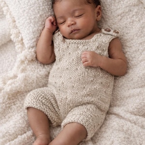 May include: A beige knitted romper featuring a textured stitch pattern, a sleeveless design, and two small brown buttons on the shoulder straps. The garment is crafted from a soft, light-colored yarn and provides a simple, functional fit for an infant.