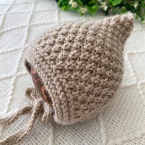 May include: A beige knitted baby bonnet with a pointed crown and tie strings. The bonnet is made of a textured knit fabric and is displayed on a white, patterned surface. The bonnet is designed to keep a baby's head warm.