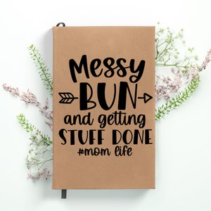 May include: A brown leather journal with the text "Messy Bun and getting stuff done #mom life" in black lettering. The journal has a black arrow pointing to the right and left of the word "Bun".