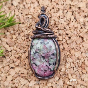 Könnte beinhalten: Handgefertigter Anhänger mit einem polierten ovalen Stein in Rosa- und Grüntönen, eingefasst in Kupferdraht. Die Drahtarbeit umfasst komplizierte Spiralen und Spulen, die ein einzigartiges, künstlerisches Design schaffen. Der Anhänger hängt an einer Kupferschlaufe.