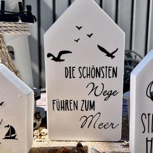 May include: White ceramic house-shaped sign with black text that reads "Die sch&ouml;nsten Wege f&uuml;hren zum Meer" (The most beautiful paths lead to the sea). There are also black silhouettes of seagulls flying above the text.