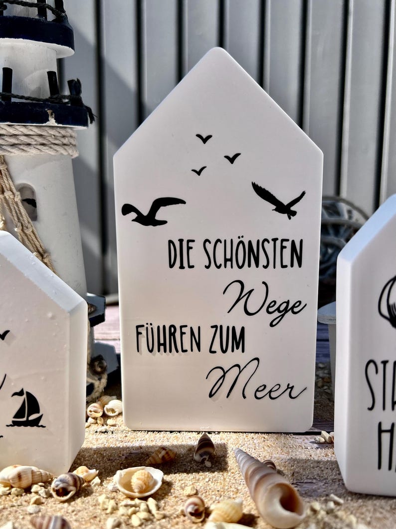 May include: White ceramic house-shaped sign with black text that reads "Die sch&ouml;nsten Wege f&uuml;hren zum Meer" (The most beautiful paths lead to the sea). There are also black silhouettes of seagulls flying above the text.