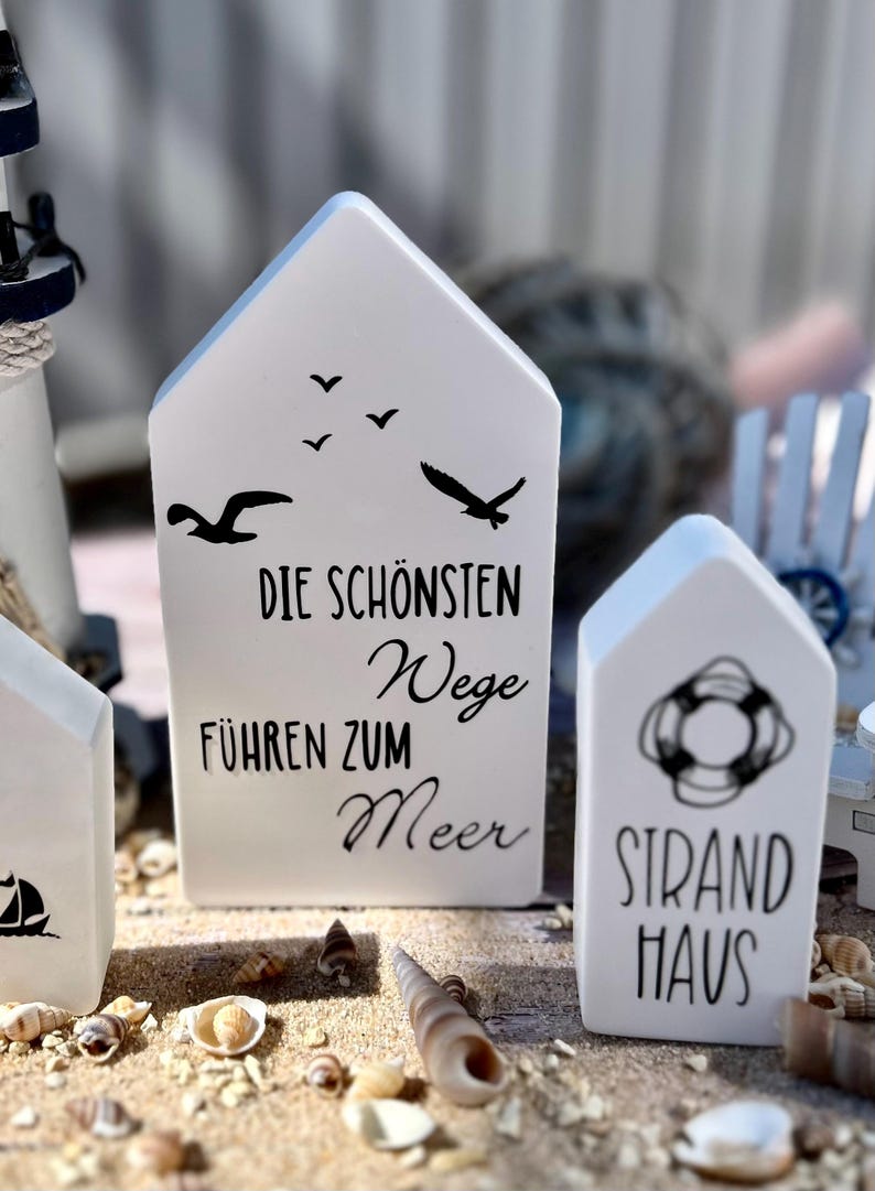 May include: Three white wooden house-shaped signs with black text and images. The first sign says "Die sch&ouml;nsten Wege f&uuml;hren zum Meer" with a silhouette of seagulls flying. The second sign says "Strand Haus" with a life preserver image. The signs are sitting on a sandy surface with seashells.