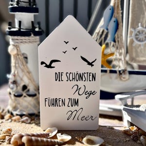 May include: White wooden sign with black text that reads "Die sch&ouml;nsten Wege f&uuml;hren zum Meer" (The most beautiful paths lead to the sea). The sign is decorated with silhouettes of seagulls.
