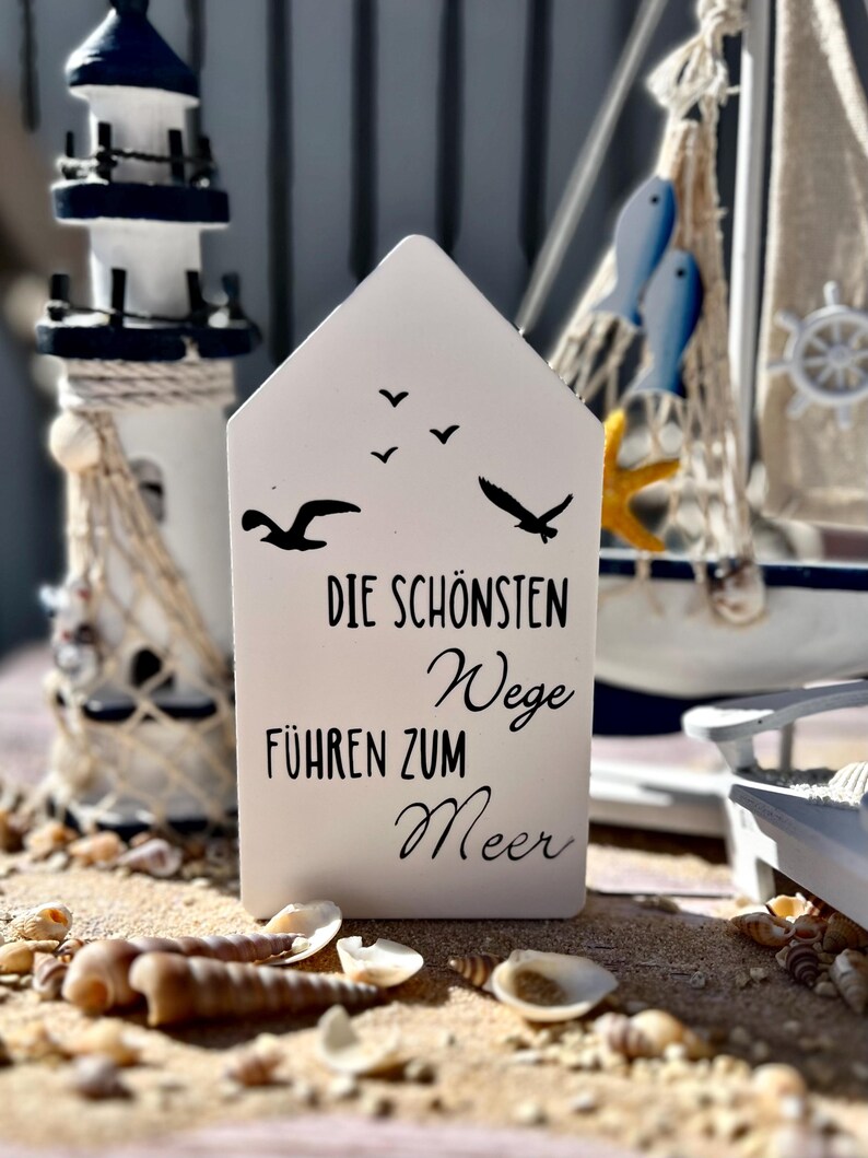 May include: White wooden sign with black text that reads "Die sch&ouml;nsten Wege f&uuml;hren zum Meer" (The most beautiful paths lead to the sea). The sign is decorated with silhouettes of seagulls.