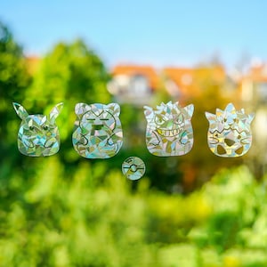 Könnte beinhalten: Eine Sammlung holografischer Fensteraufkleber mit Cartoon-Charakter-Designs. Die Aufkleber zeigen einen Pikachu, einen bärenartigen Charakter, einen Gengar und ein kleines, rundes Design. Die Designs bestehen aus schillernden, mosaikartigen Teilen, die das Licht einfangen.