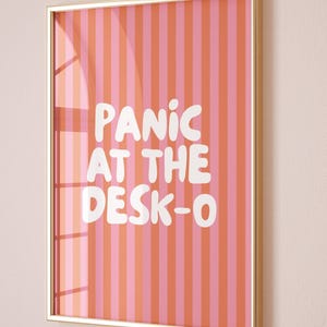 May include: Framed wall art with a gold-coloured frame. The artwork features vertical stripes in shades of pink and orange. White text reads "PANIC AT THE DESK-O". The artwork is displayed on a light pink wall.