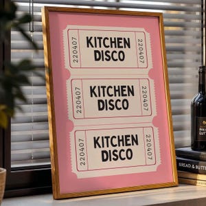 May include: A framed print with three pink and white ticket stubs, each displaying "KITCHEN DISCO" in black. The frame is light brown. The print is displayed on a windowsill, with a bottle and books visible.