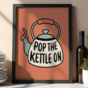 May include: A framed art print featuring a whimsical illustration of a kettle with the text "POP THE KETTLE ON" in black. The kettle is white with blue accents, set against a coral background. The print is displayed with other kitchen decor.