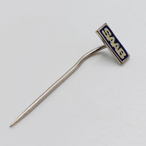 May include: A silver-toned metal lapel pin featuring a rectangular head with a dark blue enamel inlay. The word SAAB is printed in silver capital letters across the center of the blue rectangle. The pin has a long, slender, slightly curved stem.