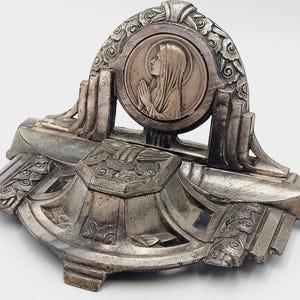 May include: A silver-toned religious shrine with an arched top featuring a circular relief of a praying figure. The base has ornate Art Deco-style detailing, including floral and geometric patterns. The piece is set against a white background.