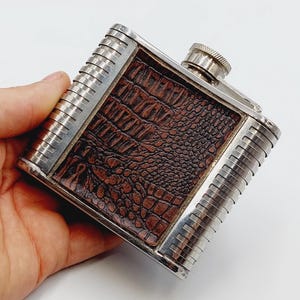 May include: A square metal flask featuring a dark brown, textured leather-like inlay with a crocodile skin pattern. The flask has a polished silver-tone finish on the sides and top, with a screw-top cap and vertical ribbed detailing on the edges.