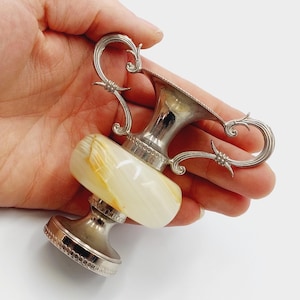 Op de afbeelding: Een zilverkleurig trofee-vormig object met een crème-kleurige, gemarmerde stenen basis. De trofee heeft sierlijke handvatten en een decoratieve rand. Het object wordt in een hand gehouden tegen een witte achtergrond.