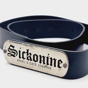 Op de afbeelding: Een zwarte riem met een rechthoekige zilverkleurige metalen plaat aan de voorzijde. De plaat toont de tekst "Sickonine" in een groot gotisch lettertype, met de tekst "ANGEL'S GATE FOREVER" direct daaronder afgedrukt.