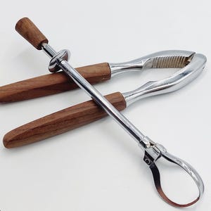 May include: A nutcracker and nut pick set. The nutcracker is silver with a ridged cracking surface and wooden handles. The nut pick is silver with a wooden handle. The set is on a white background.