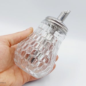 Op de afbeelding: Een glazen suikerdispenser met een zilveren deksel en tuit. De dispenser heeft een getextureerd, ruitvormig patroon.