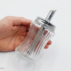 Könnte beinhalten: Ein klarer Glas-Zuckerstreuer mit silbernem Deckel und Ausgießer. Der Streuer hat ein geripptes Design und wird in einer Hand gehalten.